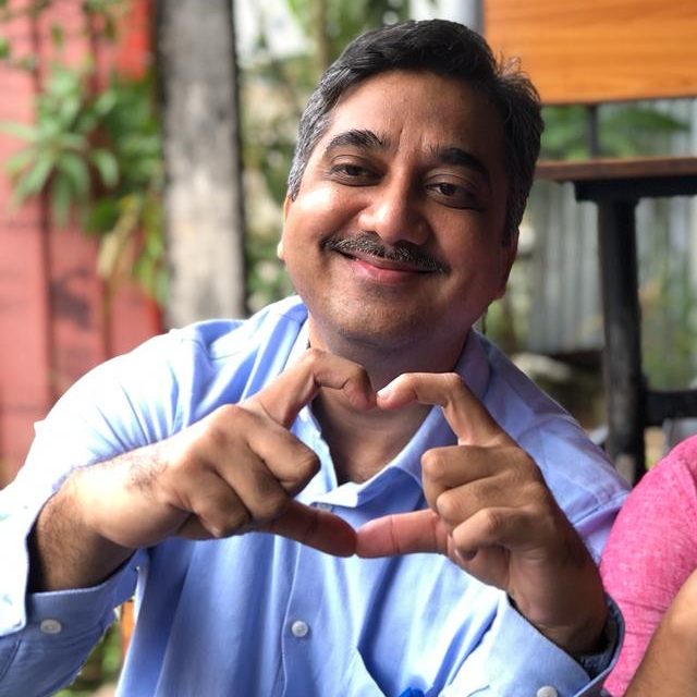 Khan Amir Maroof, a person with short, salt-and-pepper hair, a mustache, wearing a blue shirt and making a heart with his fingers