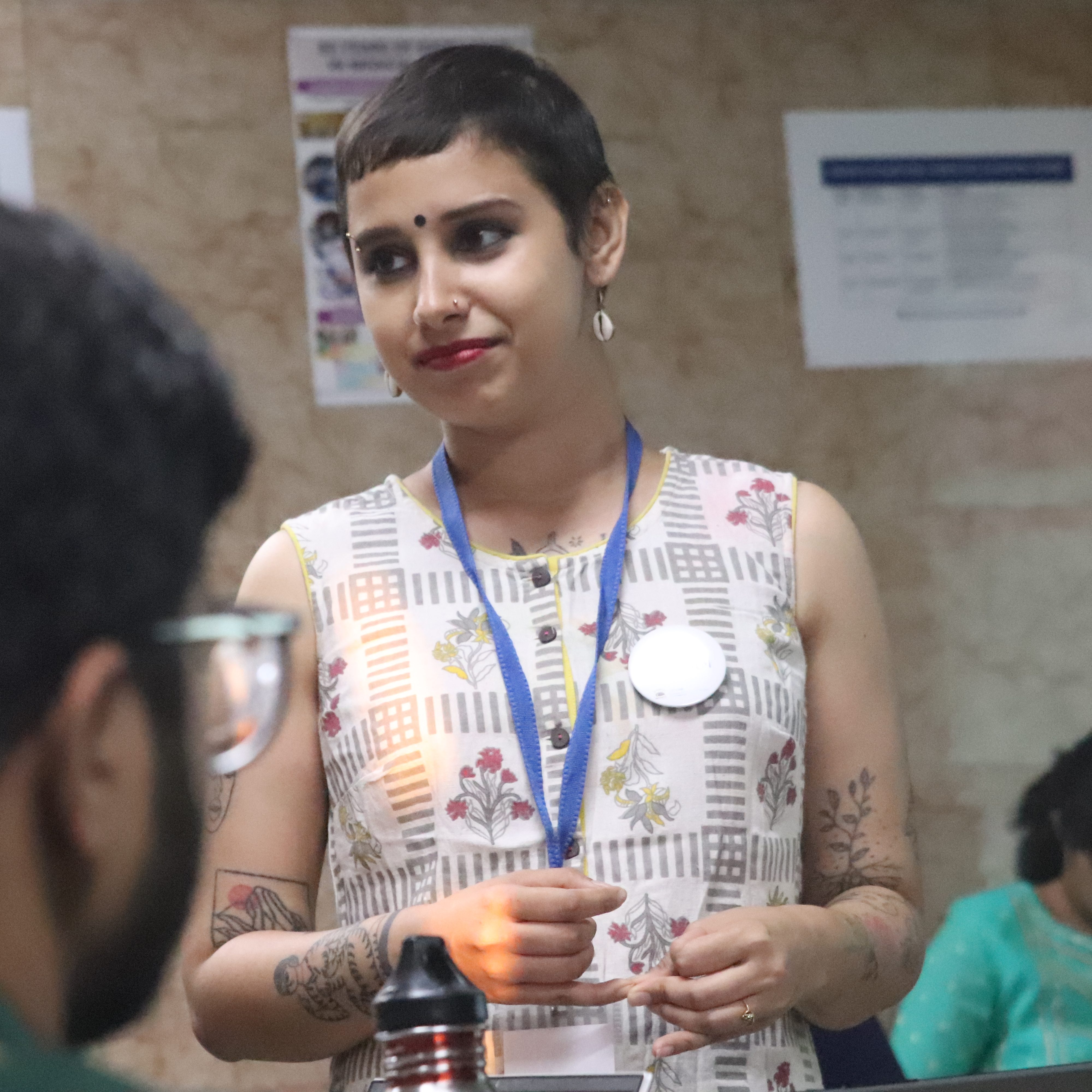 Riddhi Dastidar, a short-haired, light-skinned person with a black bindi and shell earrings, in a sleeveless patterned white kurti
