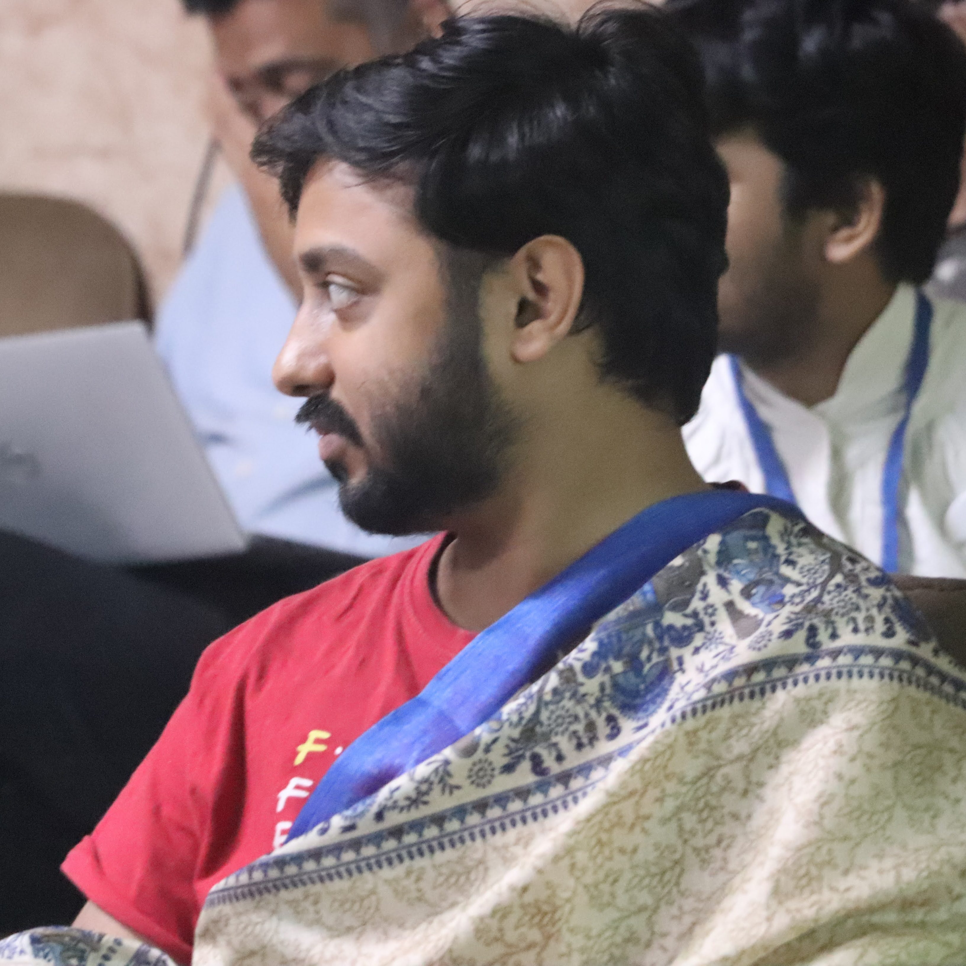 Gaurav Prateek, a queer person with short hair and a beard wearing a blue-bordered patterned sari with a red t-shirt