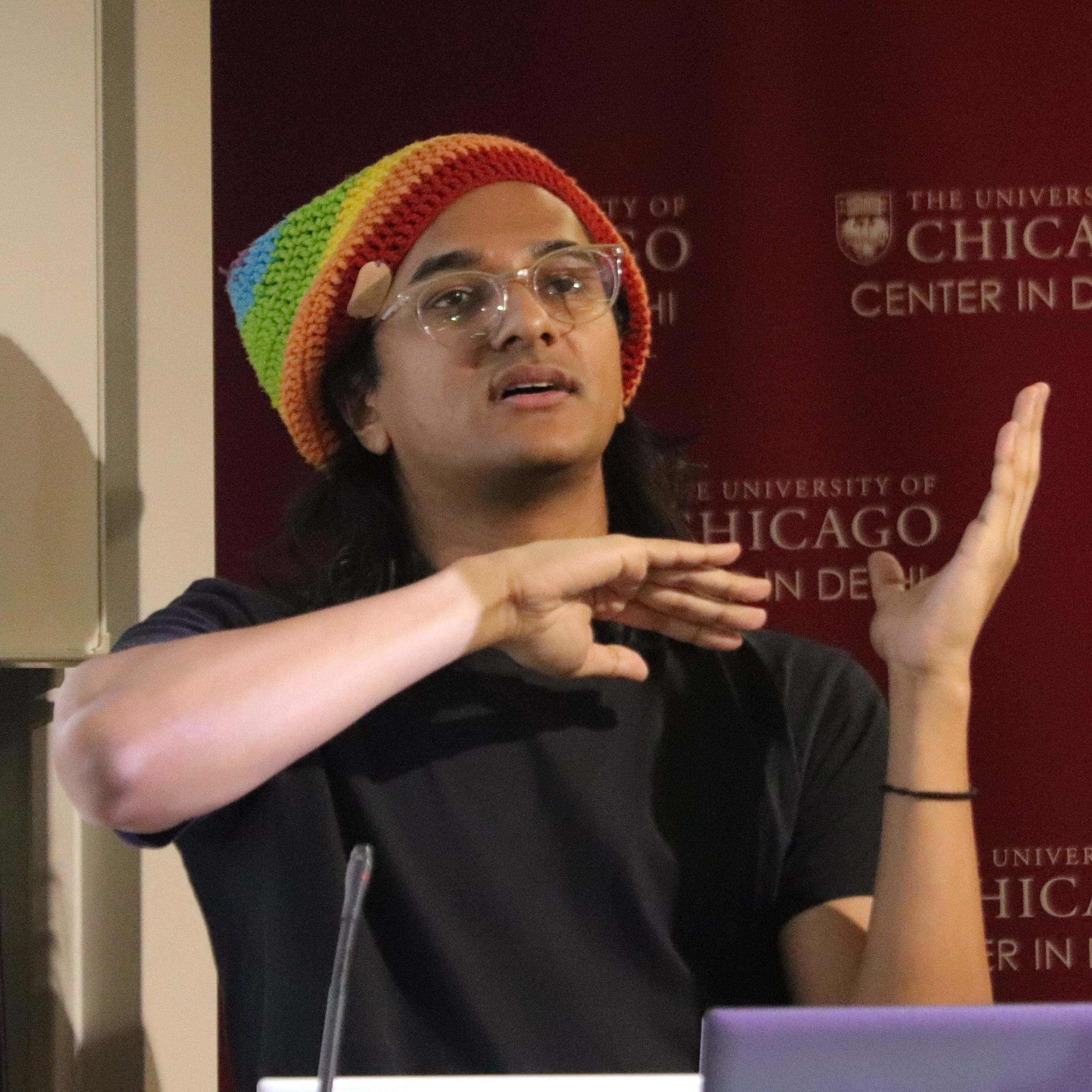 Joshua George, a queer person in a rainbow beanie speaking at a podium