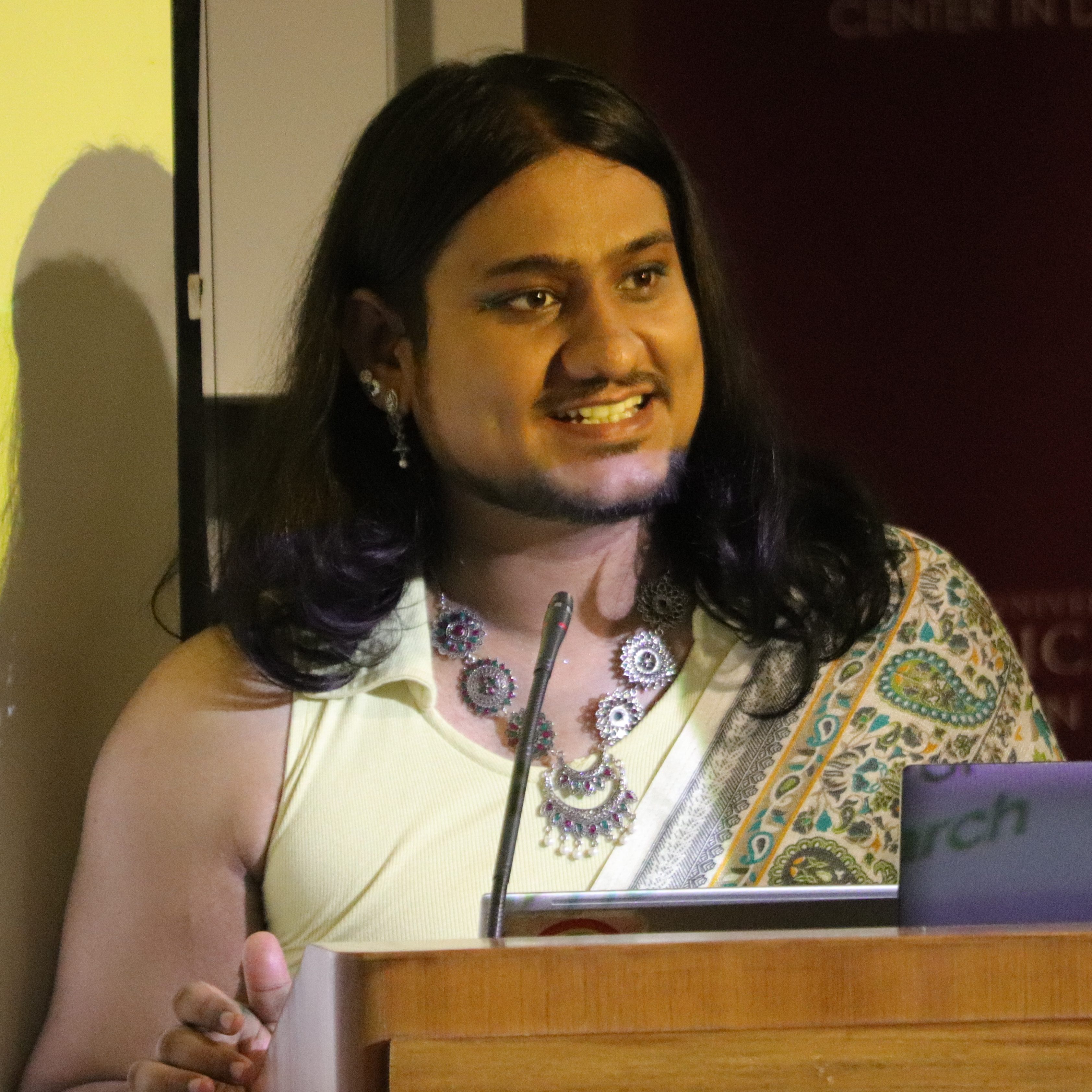 Rajan Negi, a queer person with long hair a beard, and chunky silver jewellery in a patterned green and yellow sari, with a yellow blouse, speaking at a podium