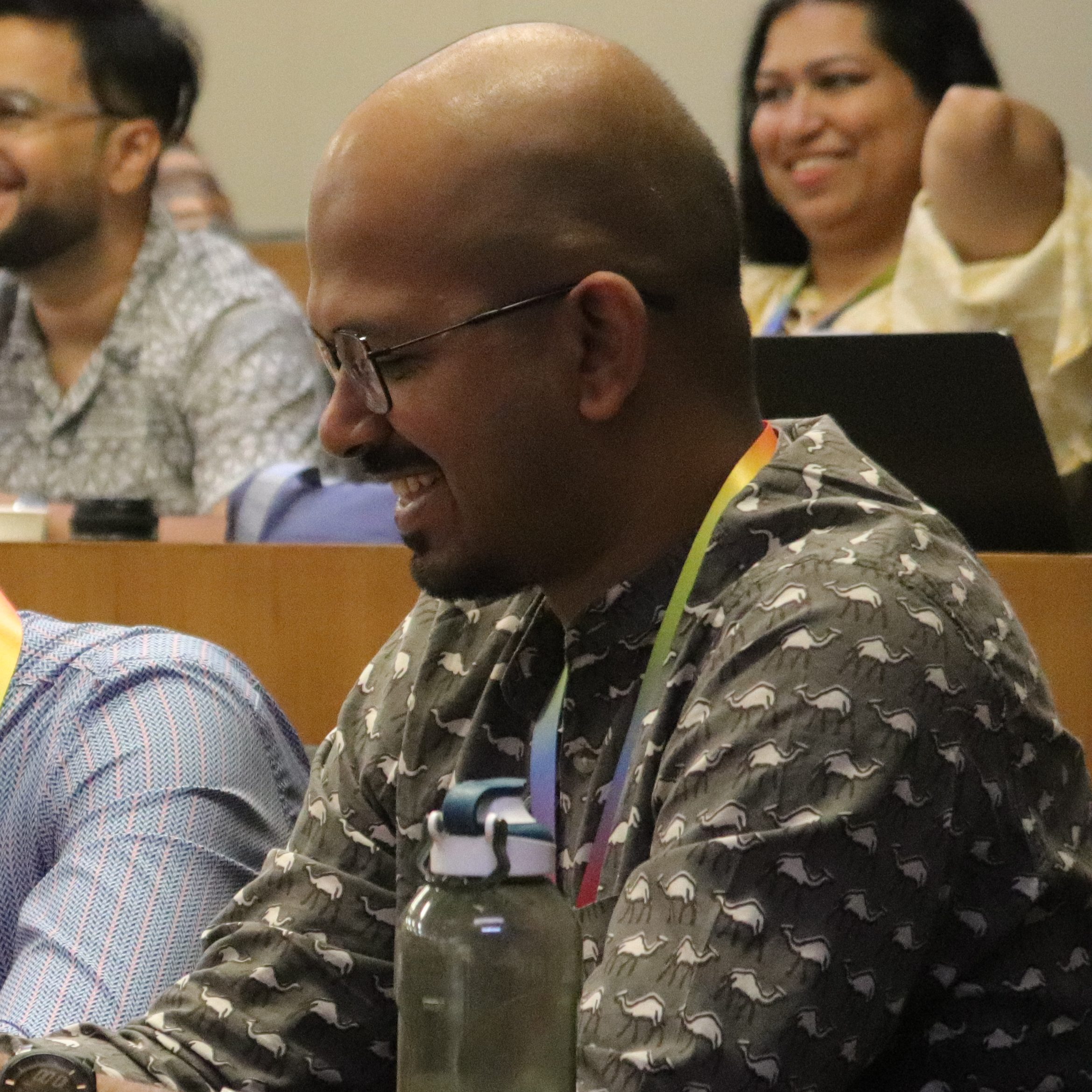 Harikeerthan Raghuram, a bald-headed person with spectacles in a grey kurta covered in camels, smiles in the audience