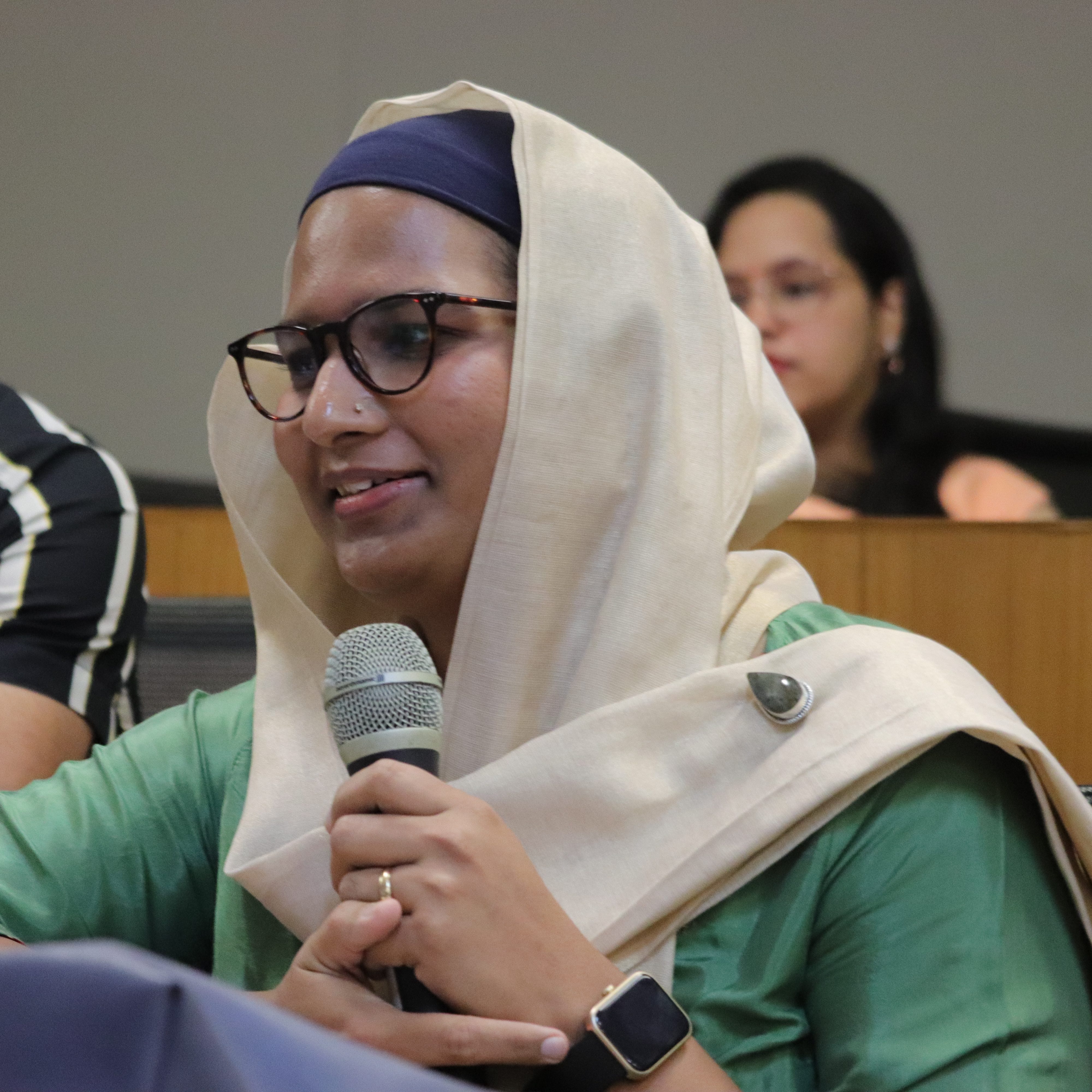 Aqsa Shaikh, a trans woman with spectacles in a beige hijab and a green kurta, speaking into a mic