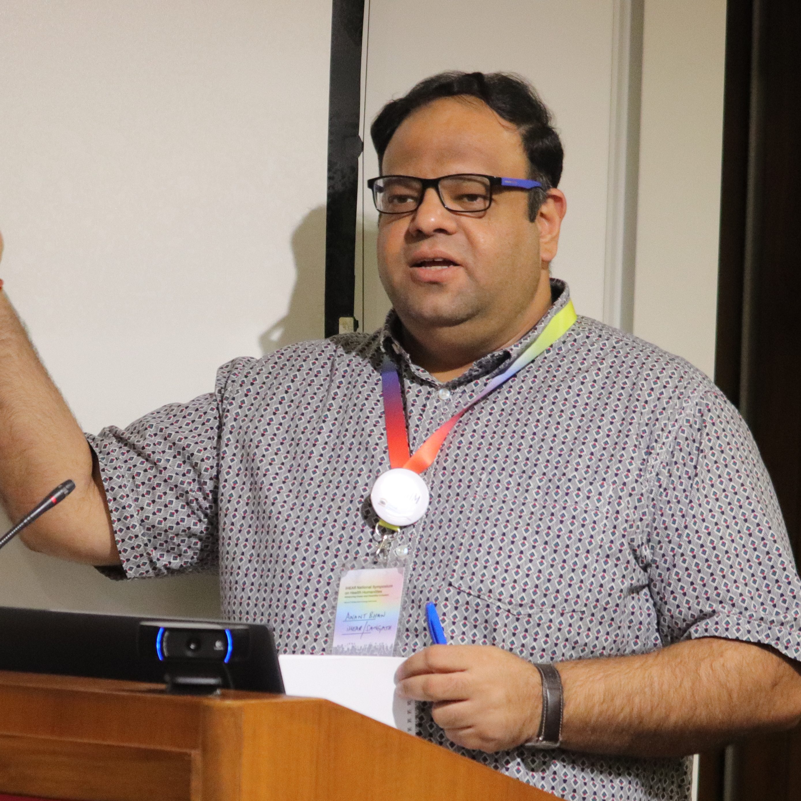 Anant Bhan, a short-haired man with rectangular glasses and a grey patterned, short-sleeved shirt, speaking at a podium