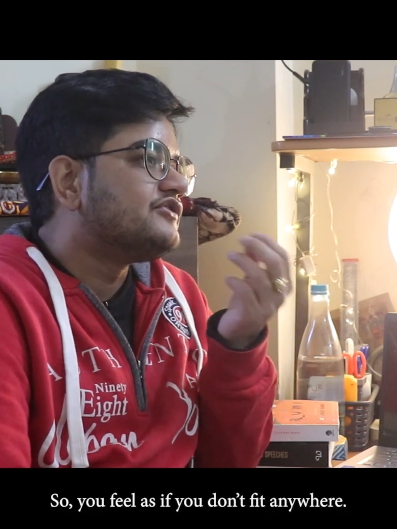 A screenshot of a video showing Satvik, a trans masc person with short hair, wearing glasses, from a side angle, wearing a red hoodie. The caption beneath him reads "So, you feel as if you don't fit anywhere."