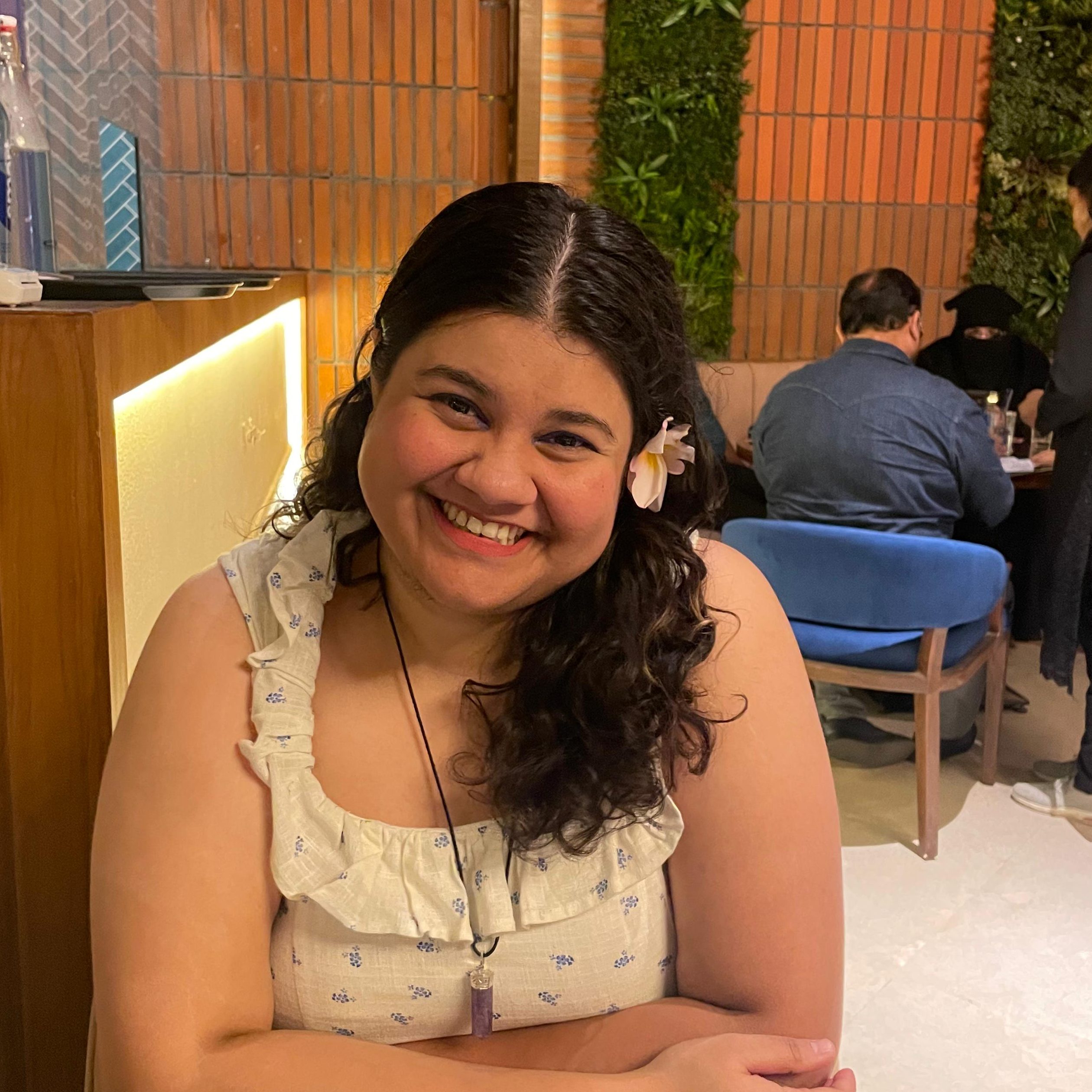 Priyanka Chakrabarty, a person with long curly hair and a flower in a hair, wearing a low-cut sleeveless white dress, smiling at the camera