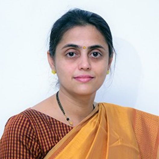Kirtana Nayak, a light-skinned person with long hair tied behind her, wearing a yellow silk sari and brown checkered blouse, with a yellow bindi on her forehead